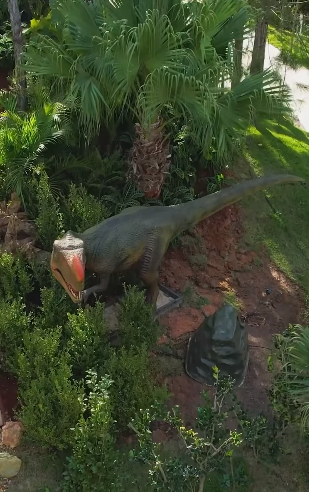 Estátua de dinossauro em um jardim com arbustos verdes e uma palmeira.