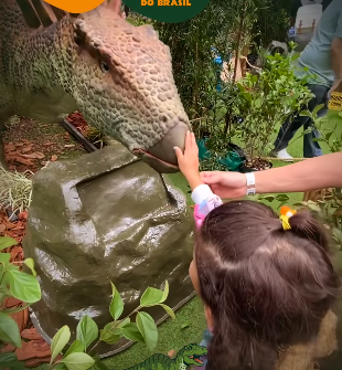 Uma criança toca o focinho de um dinossauro. O dinossauro é bege e verde. O cenário é um espaço verde ao ar livre.