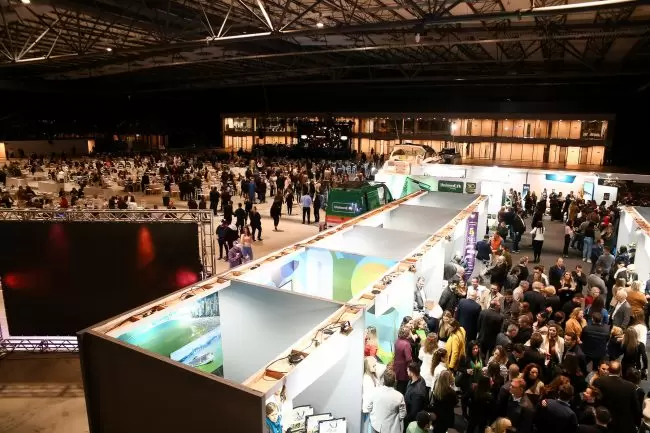 Grande evento interno com estandes de exposição, muitas pessoas interagindo e um prédio externo bem iluminado.