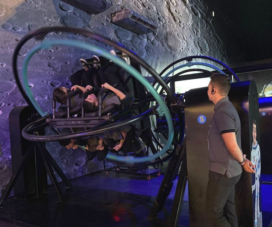 Uma pessoa opera um simulador espacial com passageiros girando dentro. Luzes azuis sobre fundo lunar.