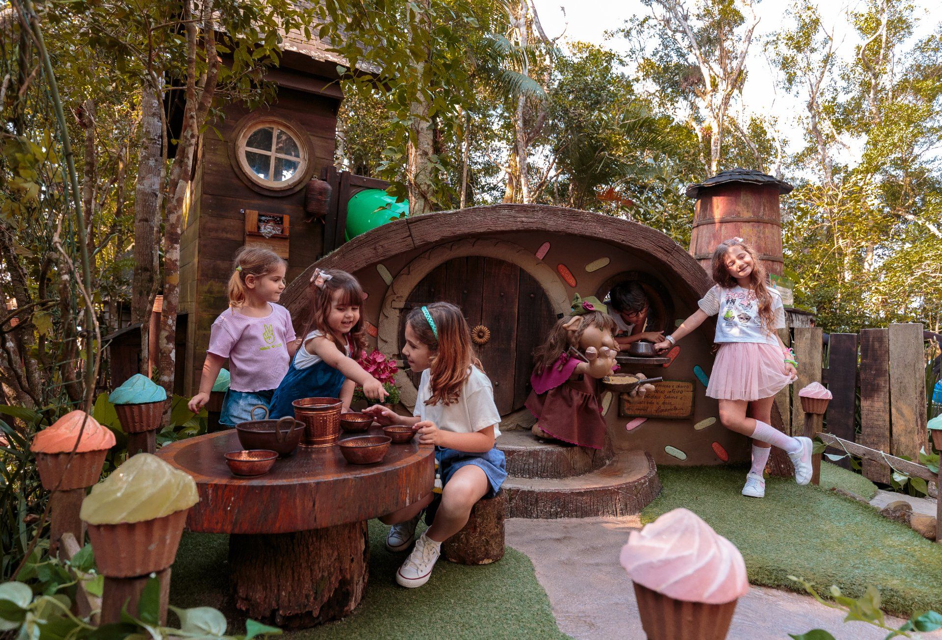 Crianças brincando em uma casa de brinquedo ao ar livre com tema de conto de fadas, decorada com cupcakes, árvores e rostos sorridentes.