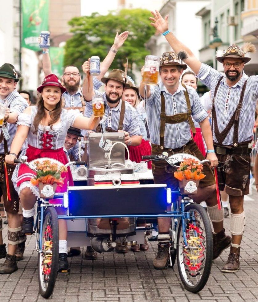 Pessoas em trajes tradicionais da Baviera em uma bicicleta de cerveja, levantando cervejas, sorrindo e comemorando na rua.