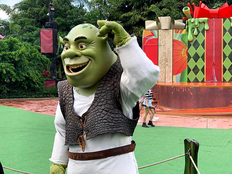 O personagem Shrek fantasiado acena e sorri em frente a um cenário com tema de presentes.