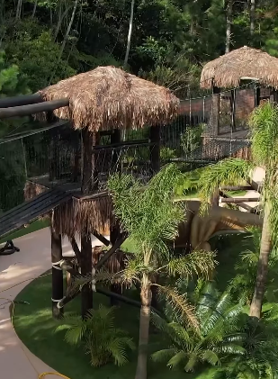 Parque infantil com estruturas de telhado de palha conectadas por uma ponte, em meio à vegetação tropical.