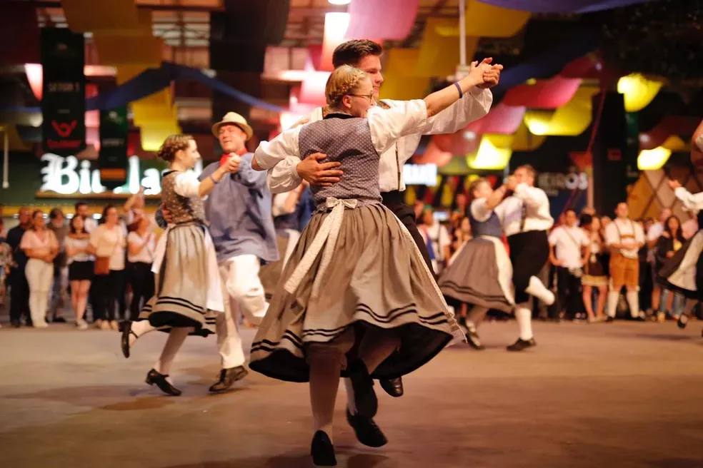 Pessoas dançando com trajes tradicionais da Baviera em um festival, ao ar livre, sob decorações coloridas.