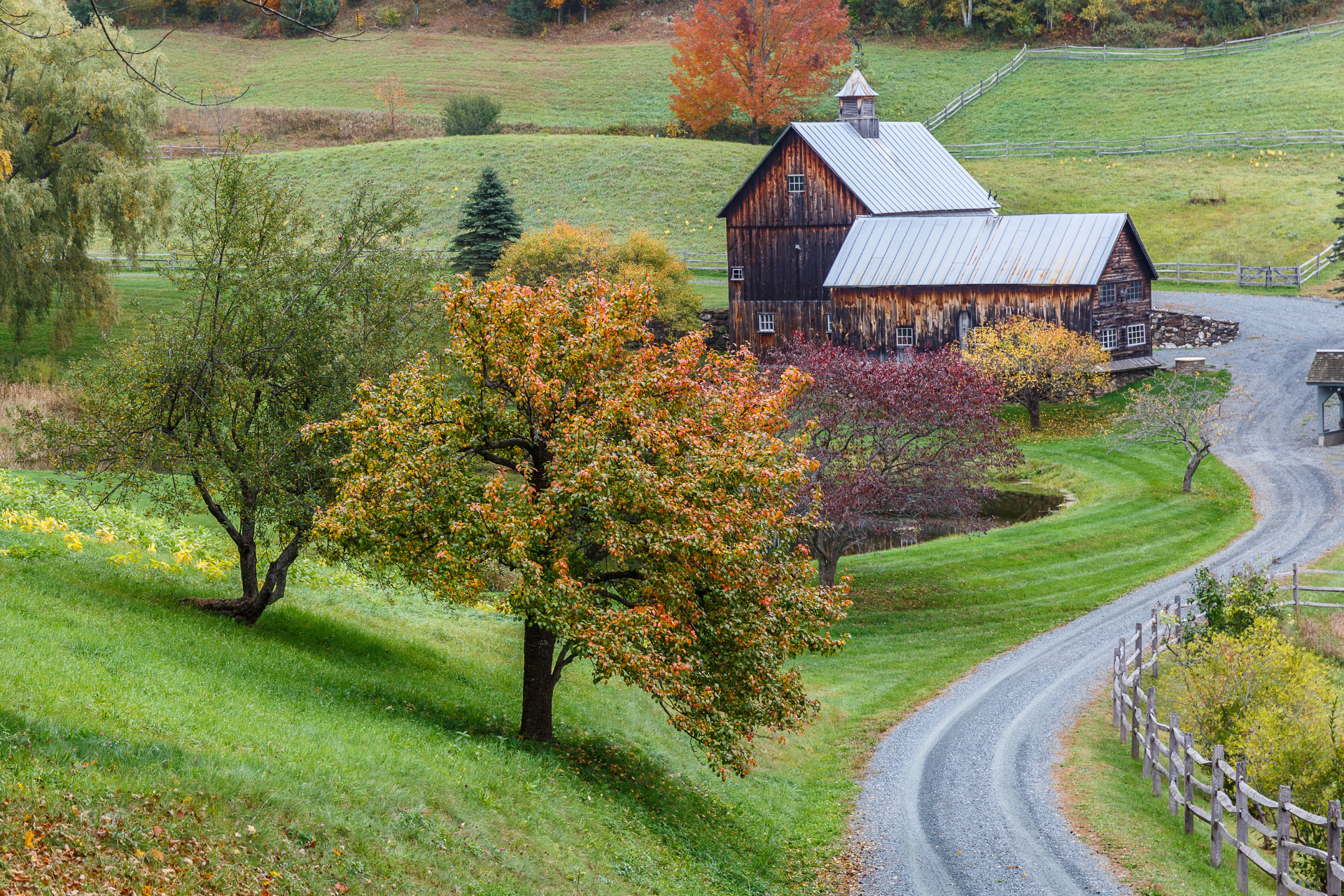 Curved country road leading past a rustic barn and autumn trees on a green hillside