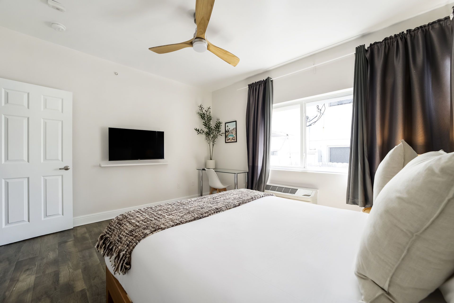 Bedroom with white walls, bed, TV, window with dark curtains, ceiling fan.