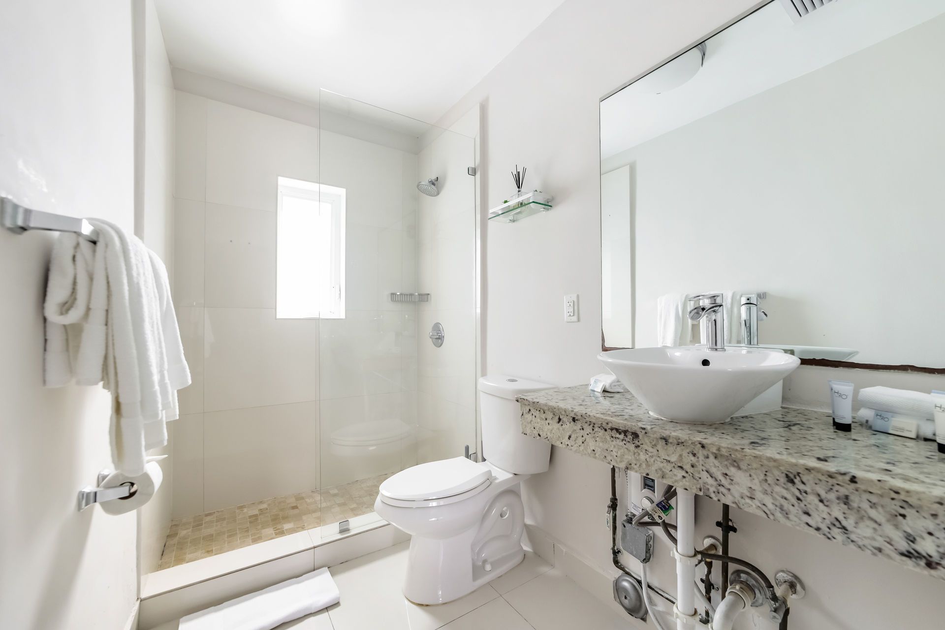 White bathroom with glass shower, toilet, sink, and granite countertop. Towel rack with white towels.