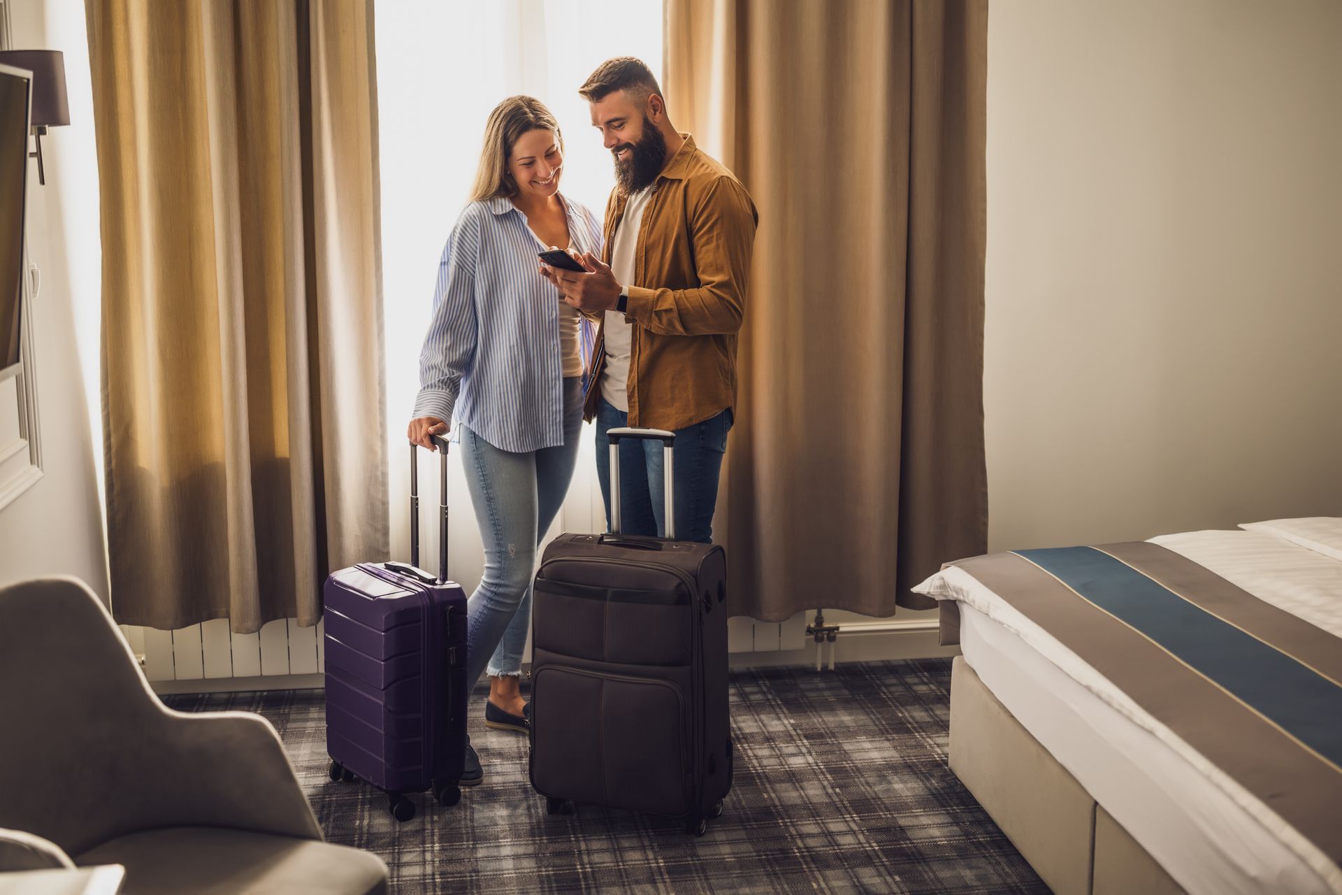 Couple in hotel room, looking at phone, with luggage.