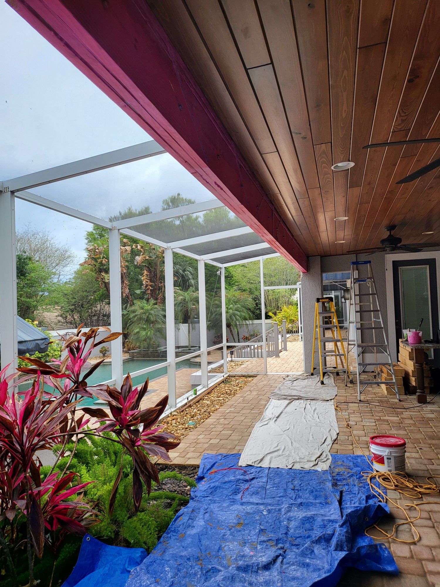 A screened-in patio with pool view. Features red trim, wood ceiling, white screens, brick flooring, and blue tarp.