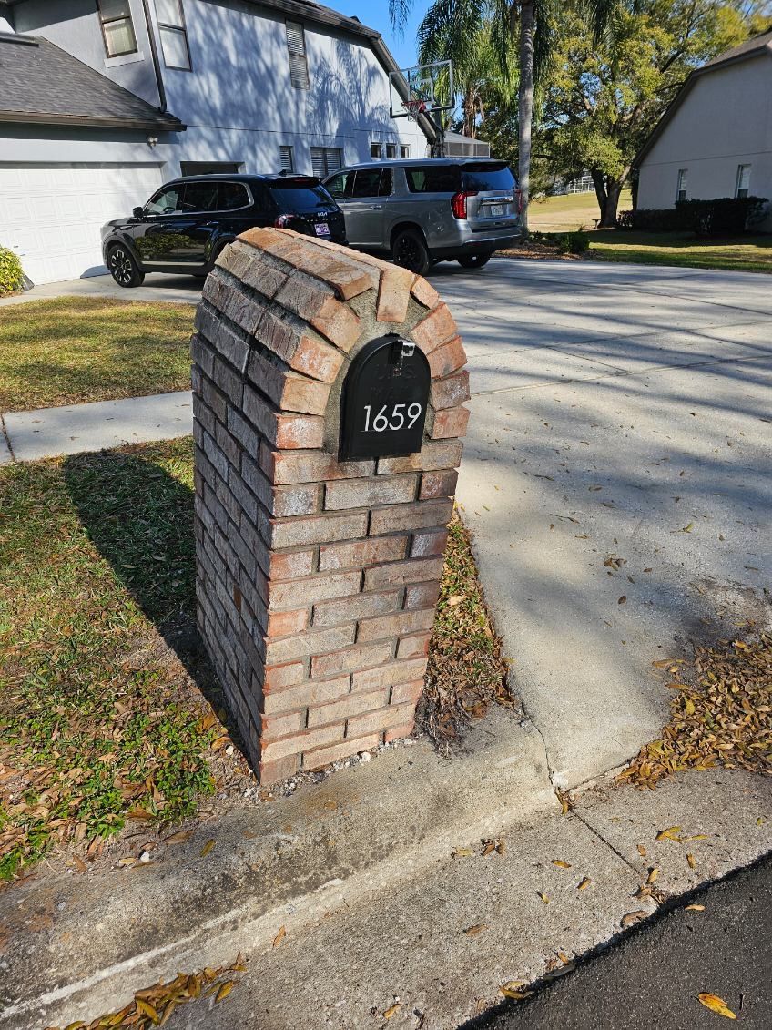 Brick mailbox with address 1659, set next to a sidewalk and street, with parked cars in the background.