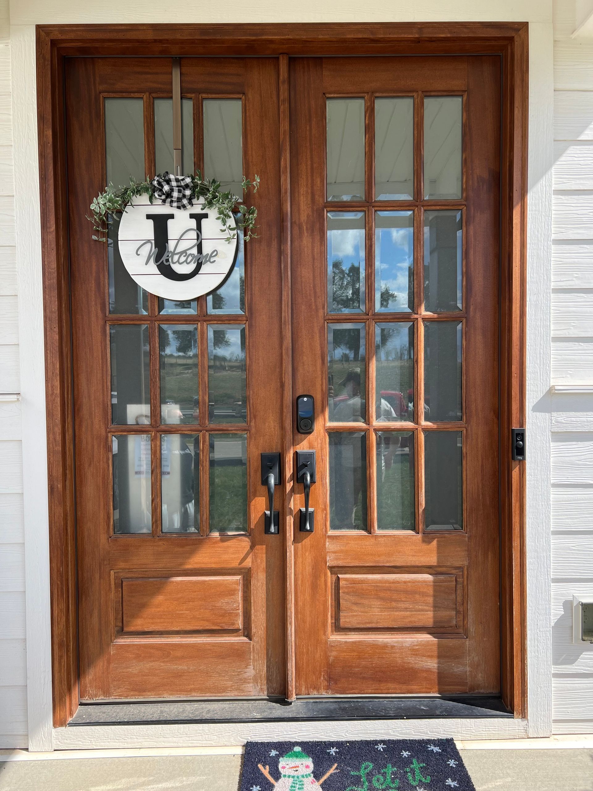 Wooden double doors with glass panes, a wreath, and a welcome mat.