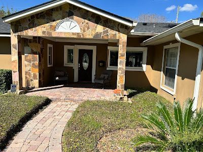 Tan stucco house with stone porch. A brick path leads to the front door. Landscaping includes bushes and a palm tree.