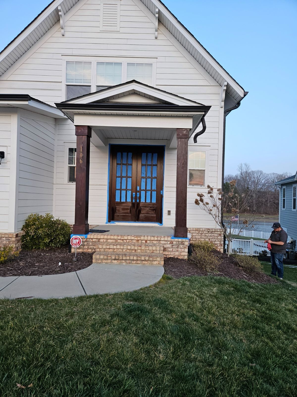 White house with dark wood pillars at the entrance, brown double doors, brick foundation, person in yard.