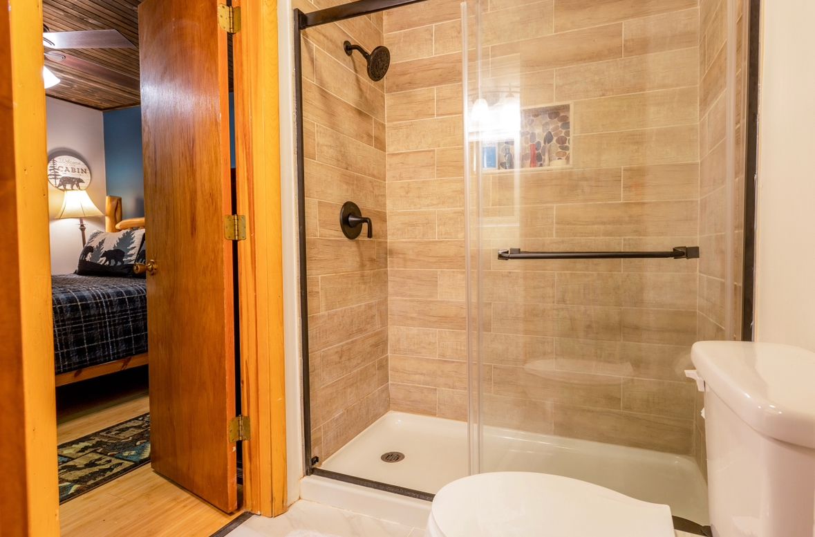 Bathroom with a glass-enclosed shower, toilet, and a doorway to a bedroom with a bed.