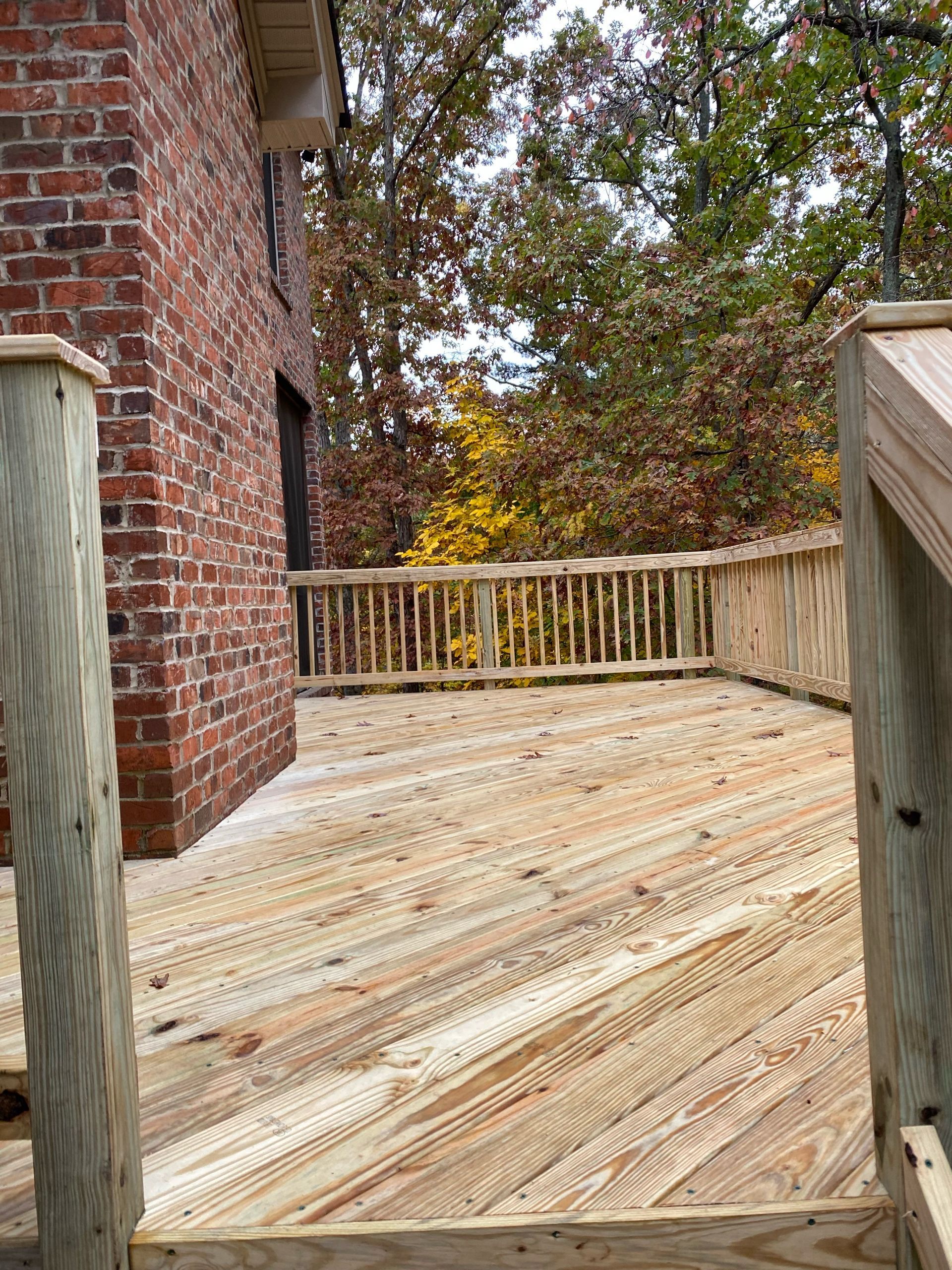 Wooden deck with railing next to a brick building. Trees with colorful leaves in the background.
