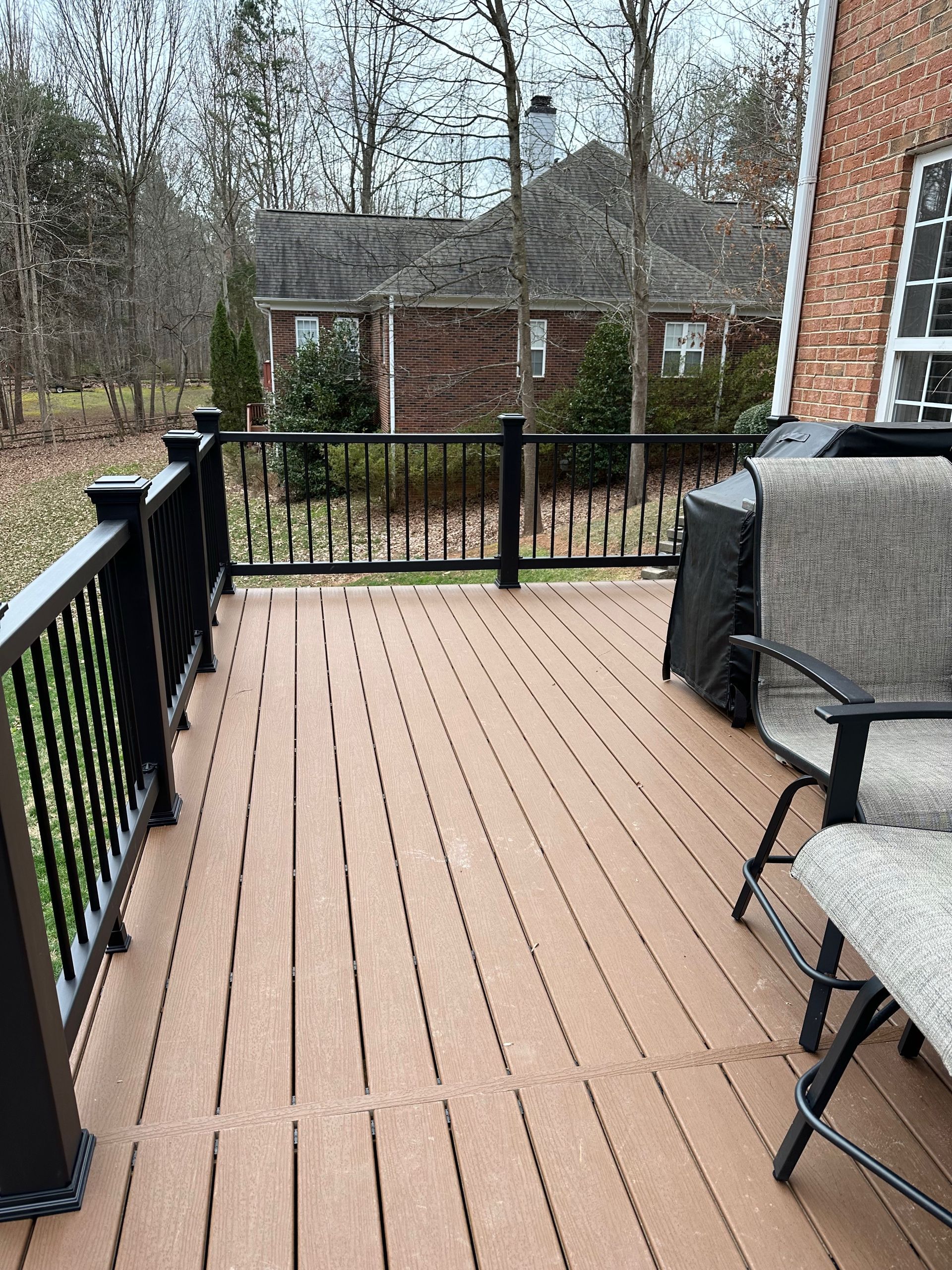 Brown composite deck with black railing and chairs; brick house in background.