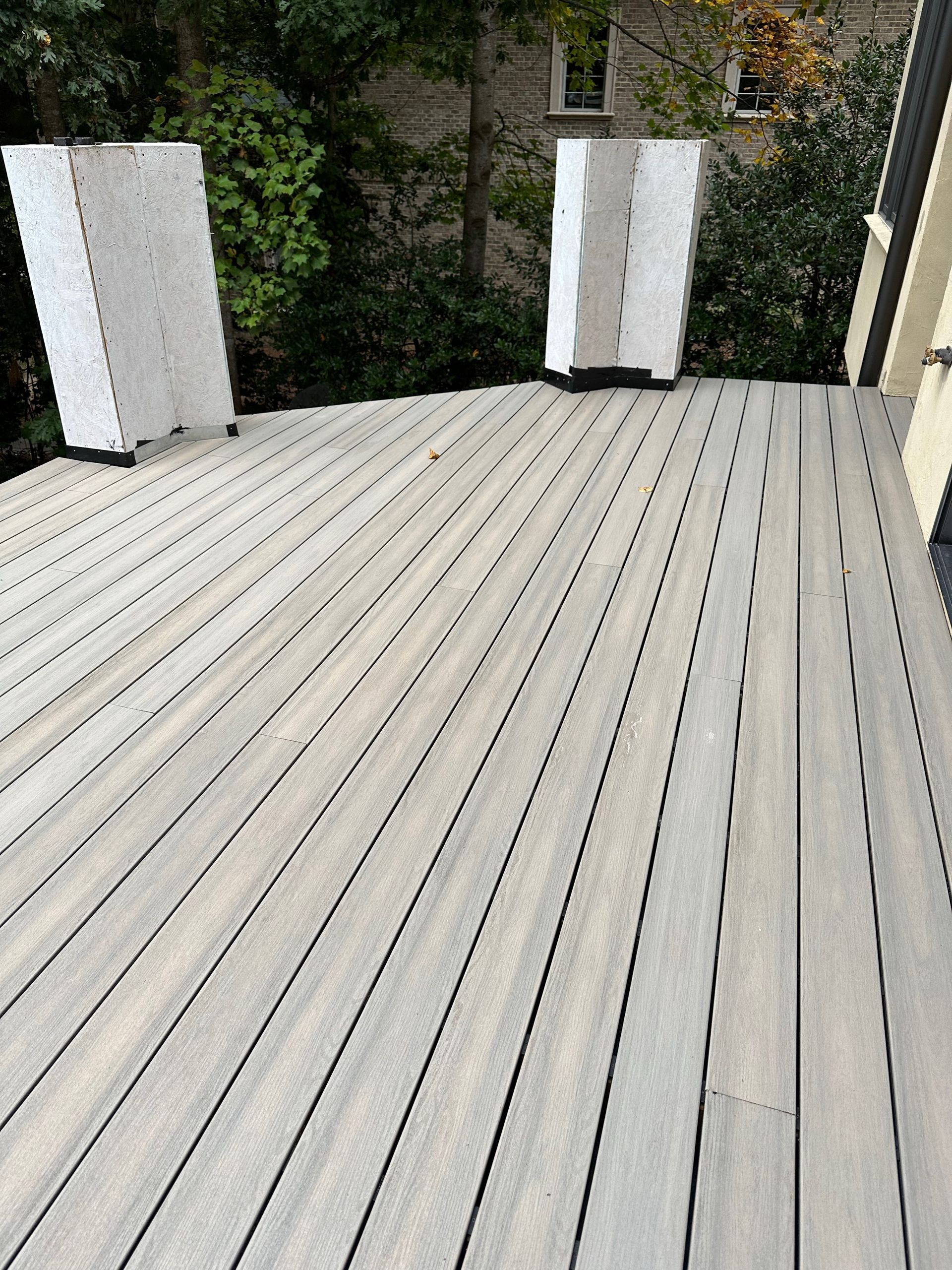 Wooden deck with light gray planks, two covered white objects, and foliage background.