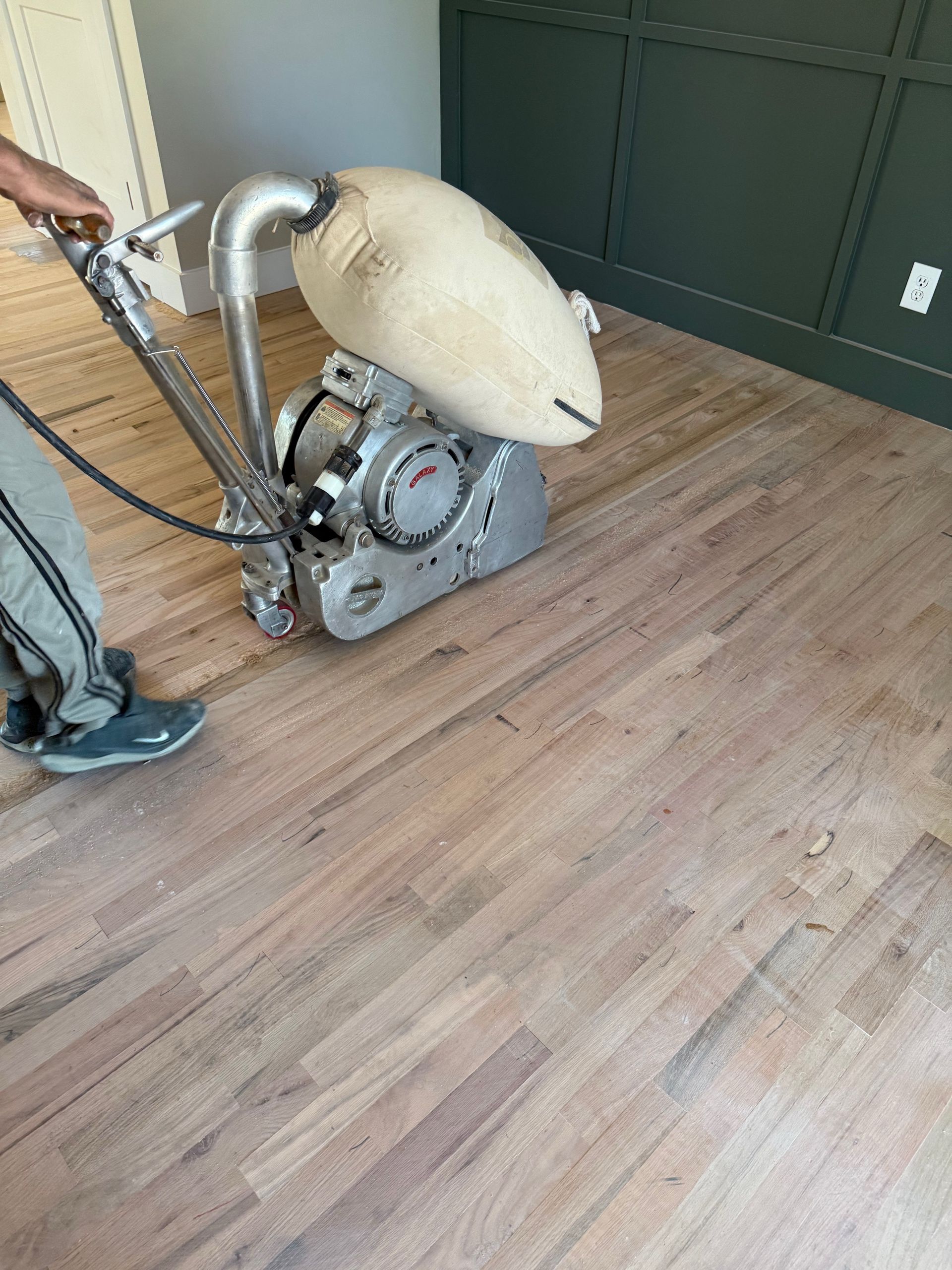 Person sanding hardwood floor with a large machine, room with green wall in background.