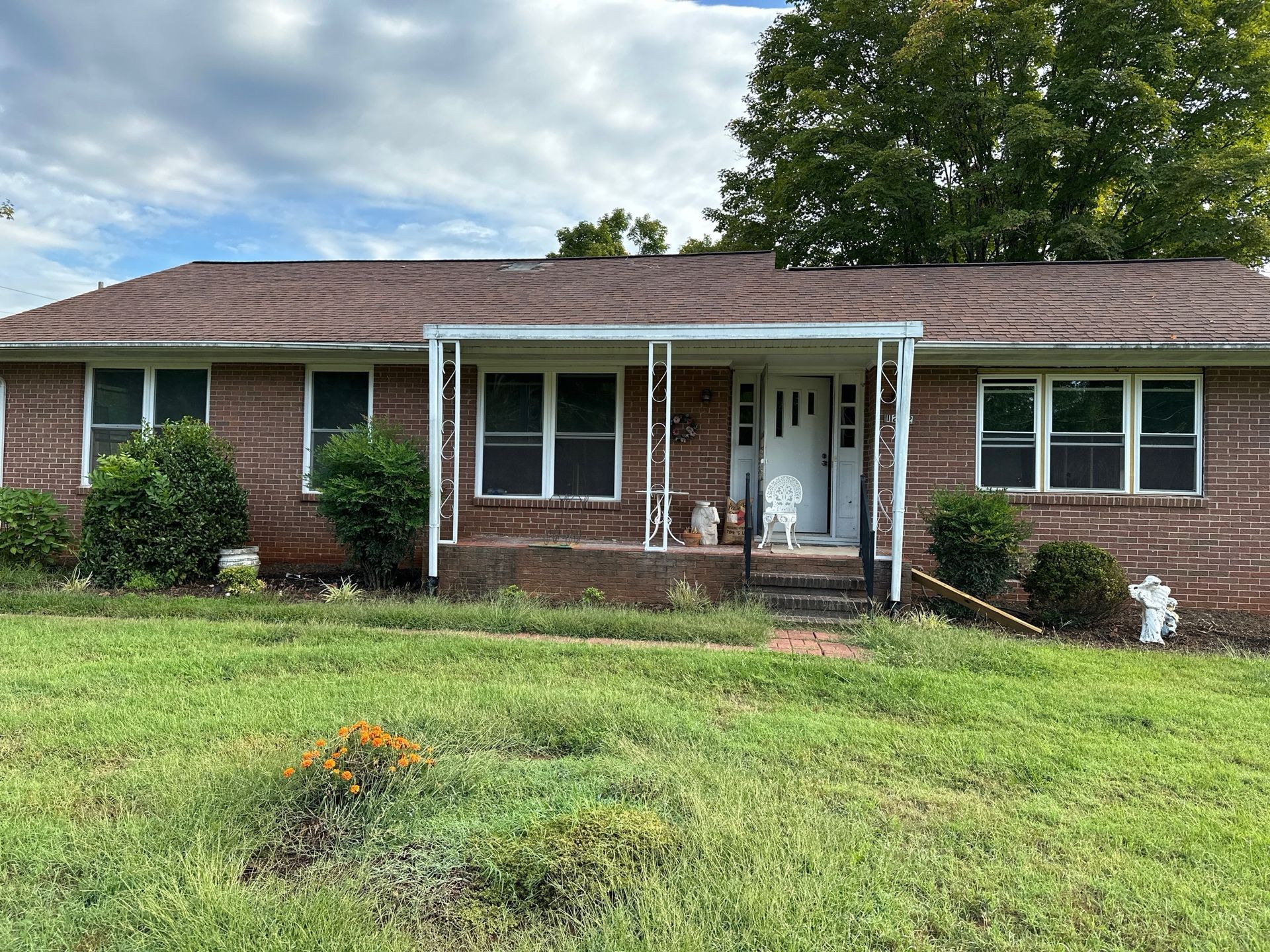 Brick house with porch and overgrown yard under a cloudy sky.