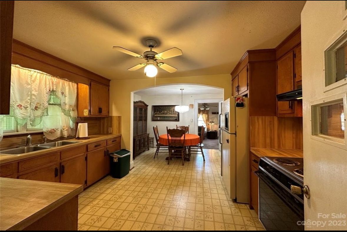 Kitchen with wooden cabinets, a view into the dining room. Cream tile floor, window with curtain.