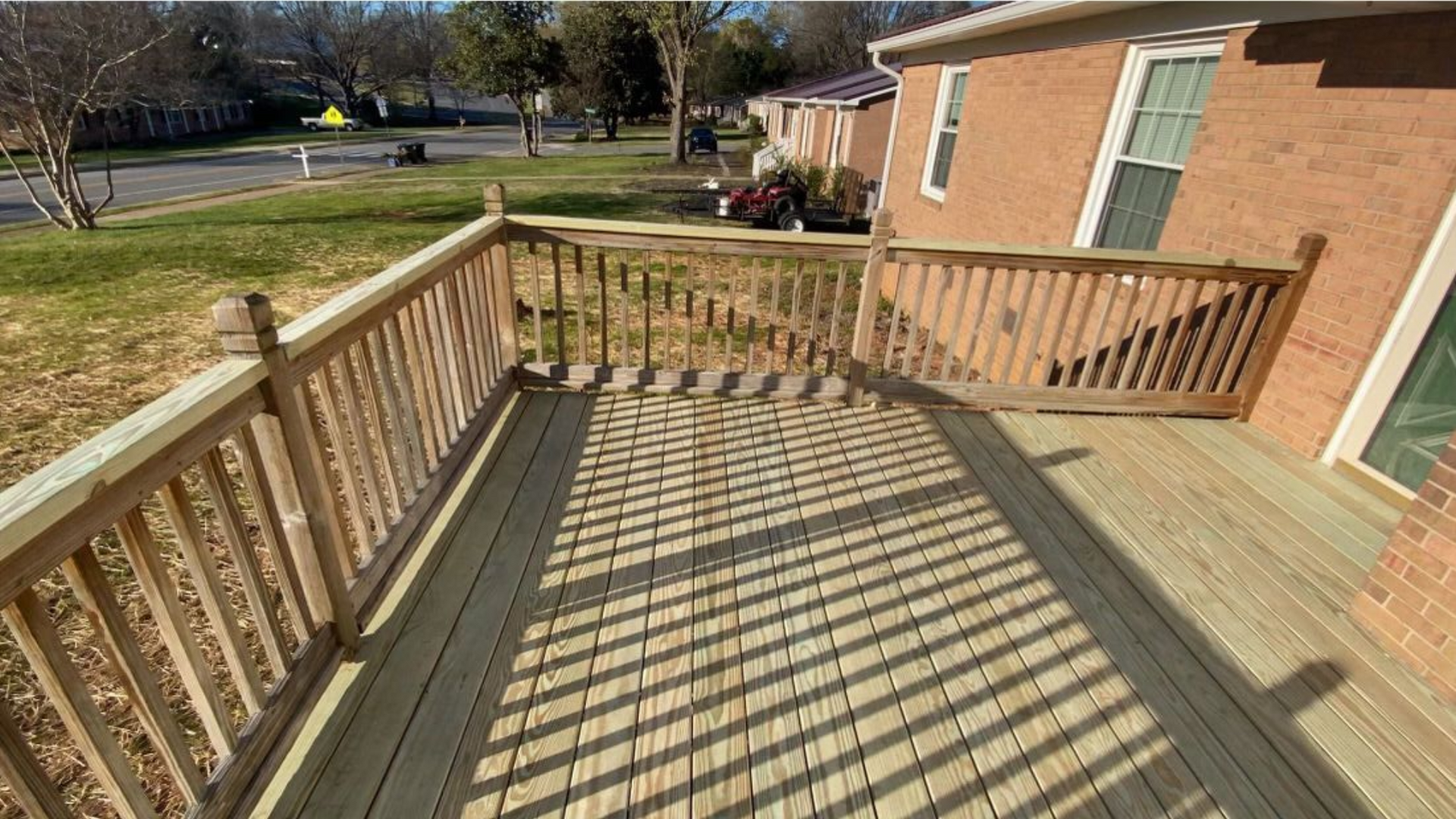 Wooden deck with railing attached to a brick house. Sunlight casts shadows on the deck boards.