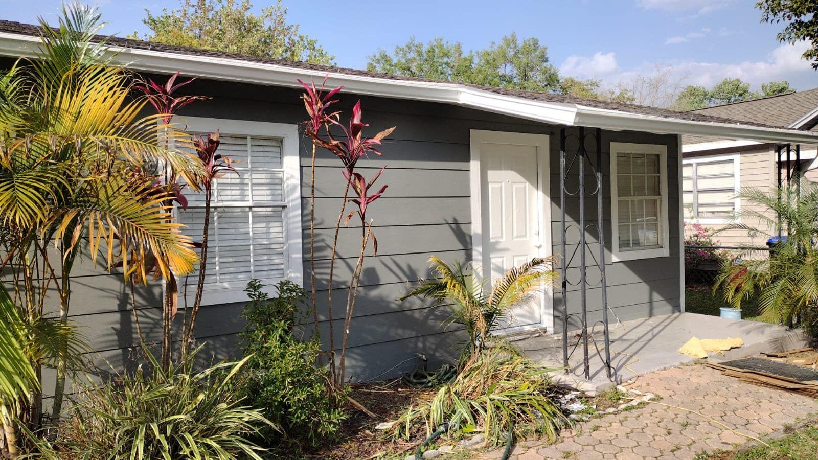 Small gray house with white trim, door, and windows. Plants in front, blue sky.