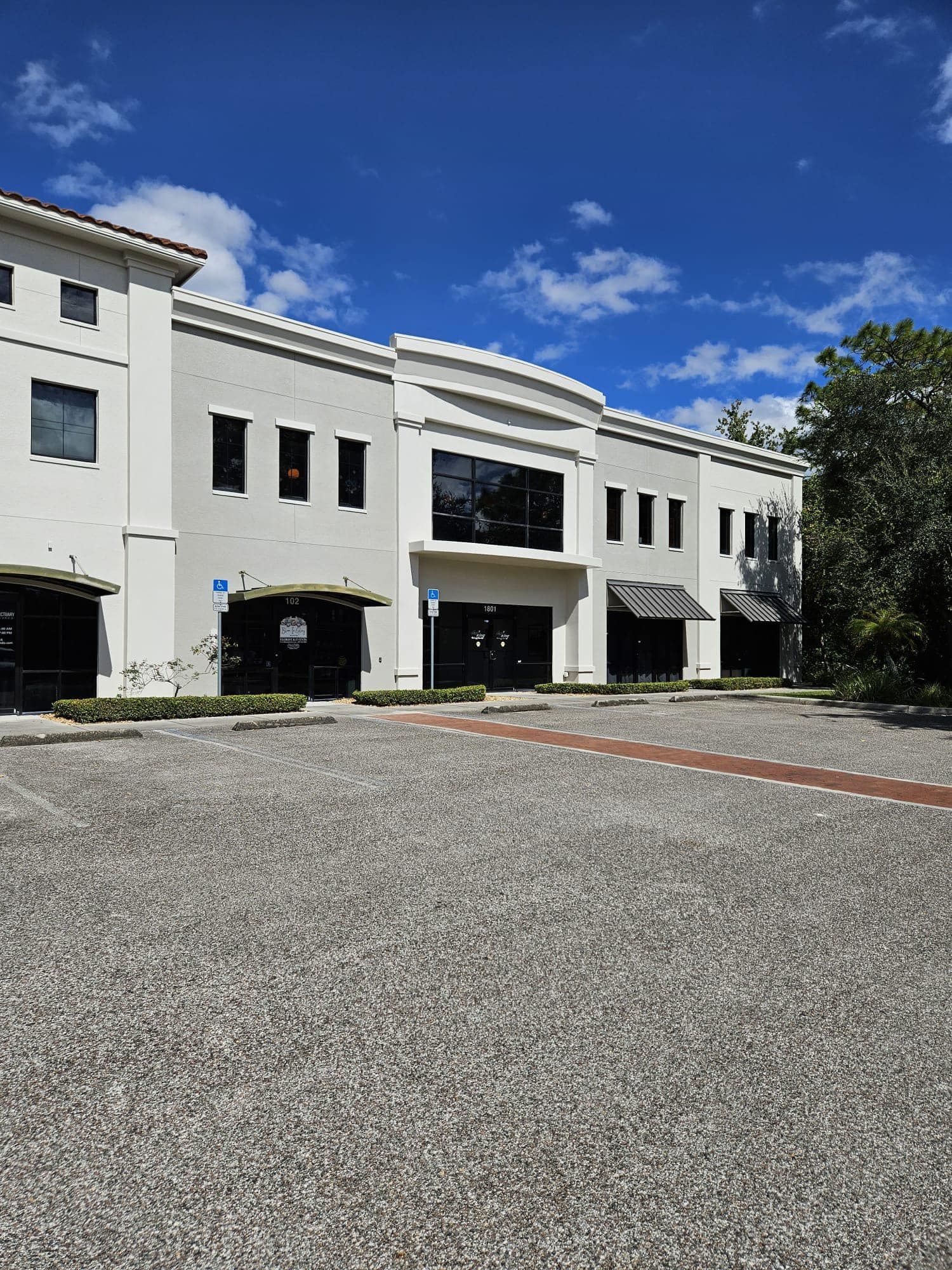 White building with dark windows and awnings, set against a blue sky, gravel parking area.