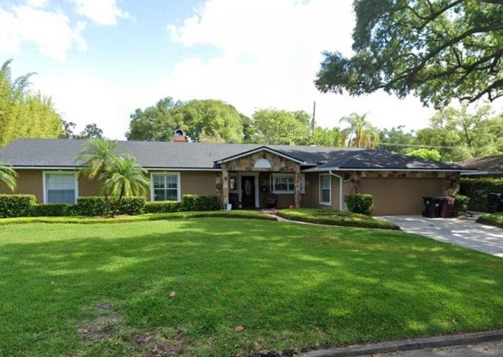 Tan house with stone facade, dark roof, green lawn, trees, and driveway.