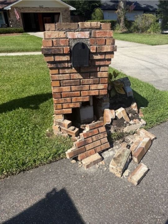 Damaged brick mailbox on a residential street; bricks are broken and fallen on the ground next to the post.