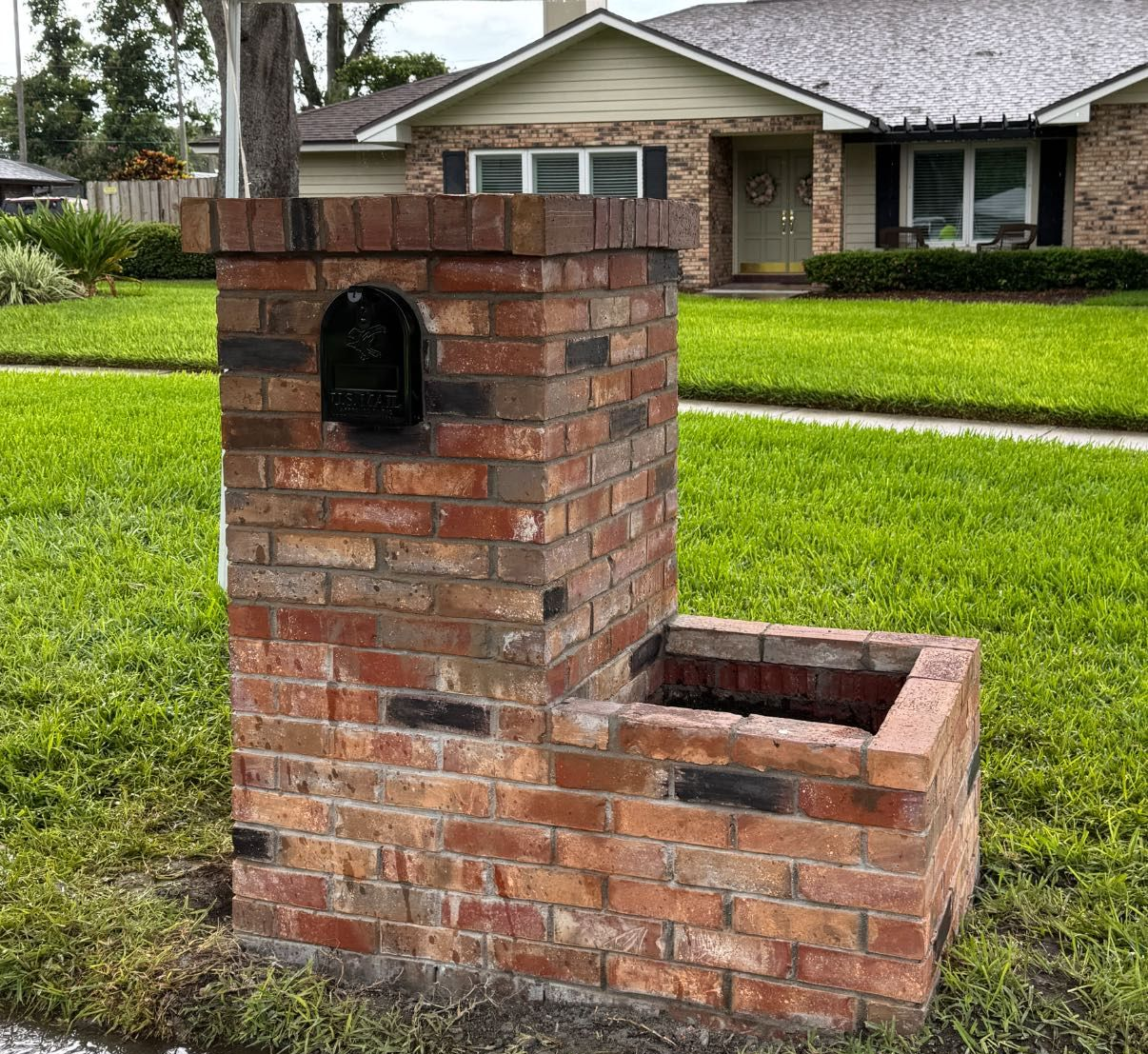 Brick mailbox with a built-in planter on a grassy lawn in front of a house.