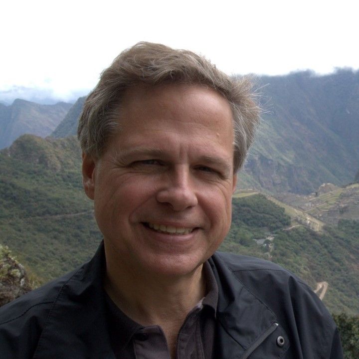 A man in a black jacket is smiling in front of a mountain range.
