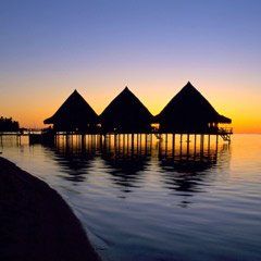 Three huts on stilts in the middle of the ocean at sunset.