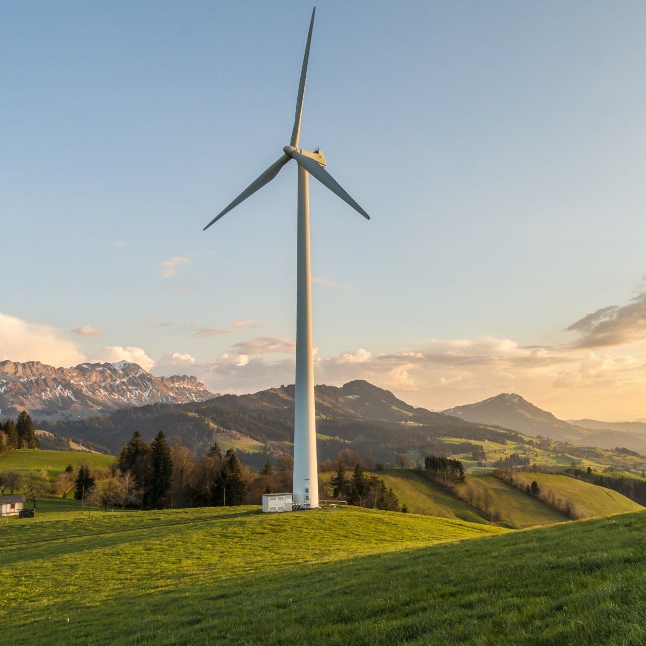 A wind turbine is sitting on top of a grassy hill with mountains in the background.
