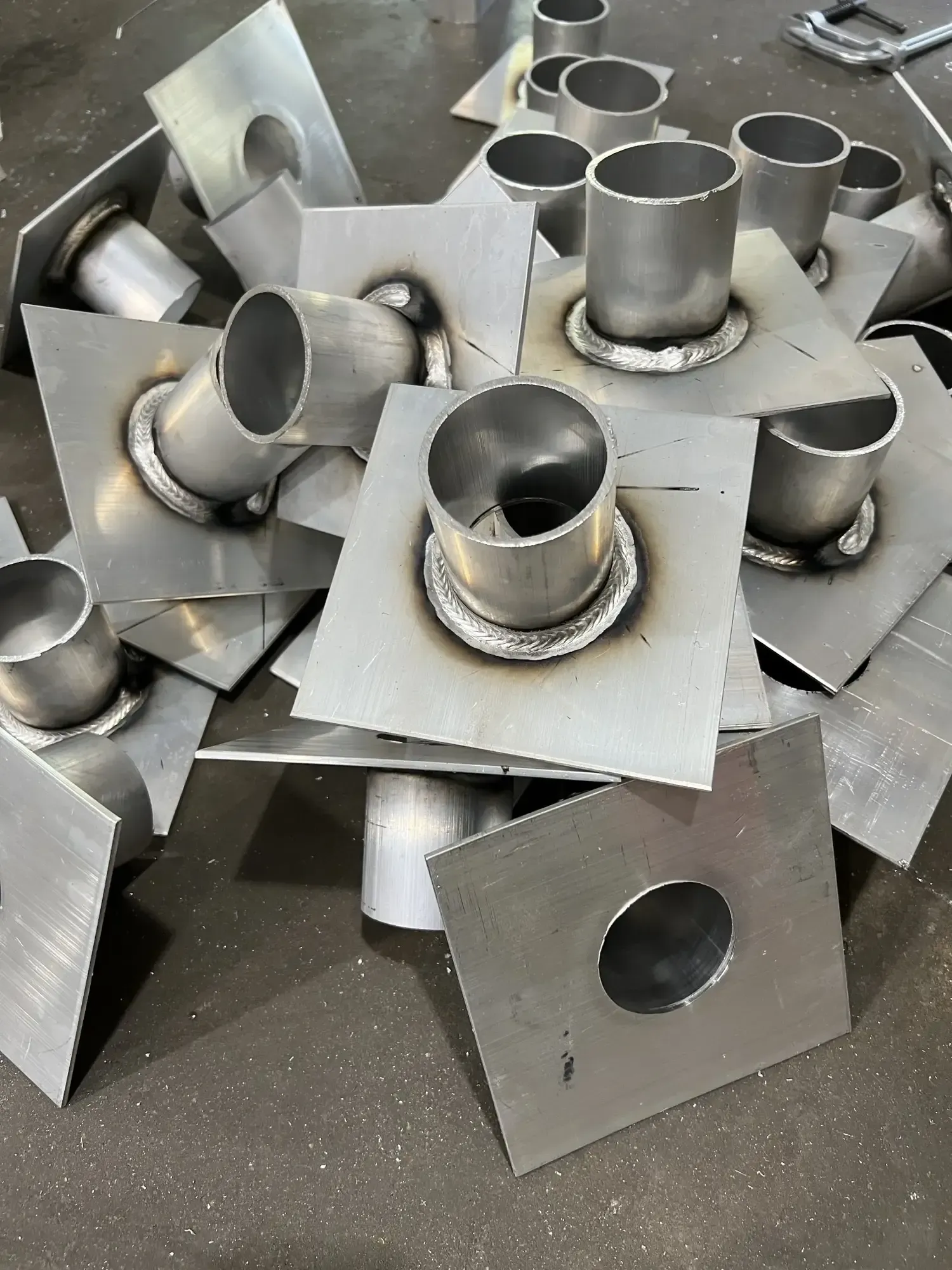 Scattered pile of welded metal parts: square plates with round tubes attached, possibly for plumbing or ventilation.