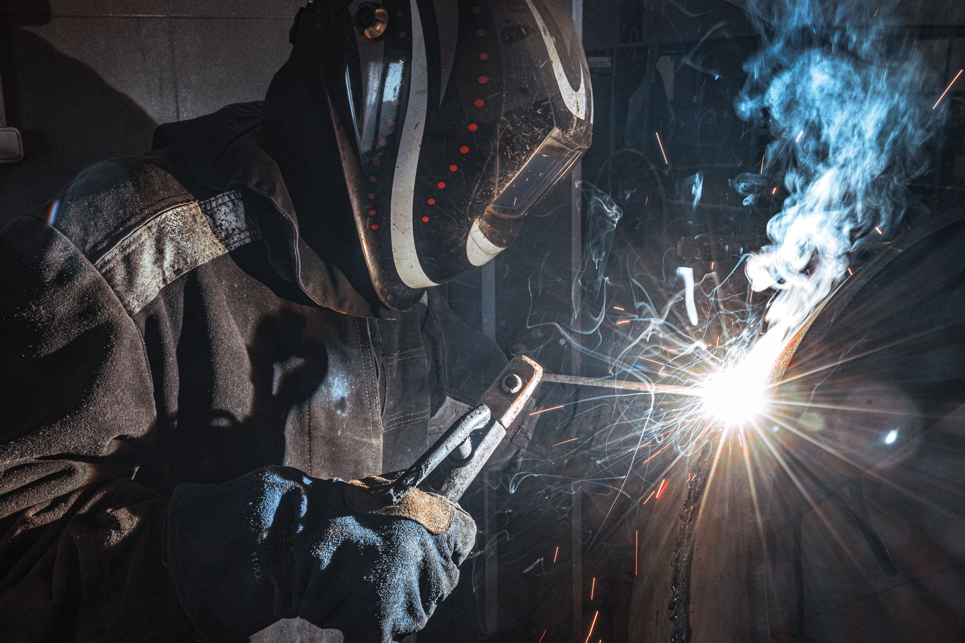 Welder wearing a protective mask welding metal, sparks and bright light in a dark setting.