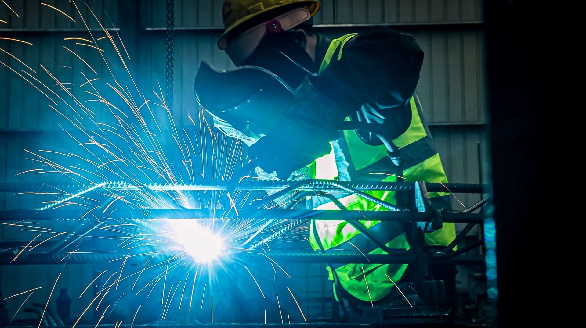 Welder wearing a safety mask and vest working on metal, with sparks flying in a dim industrial setting.