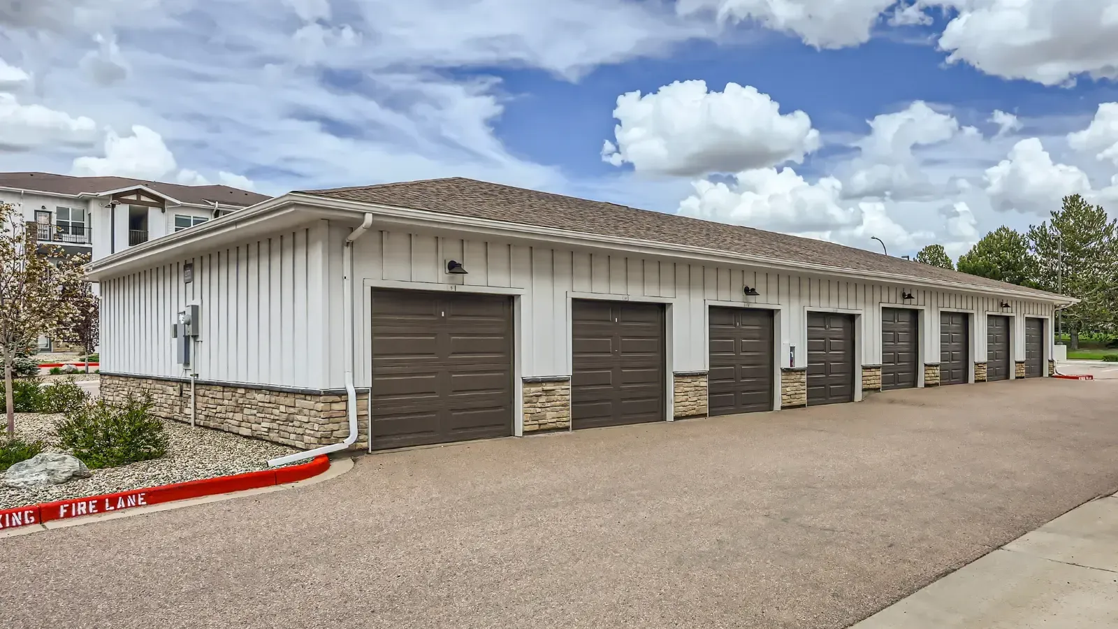 Row of six brown garage doors on a white building with stone veneer and a paved driveway.