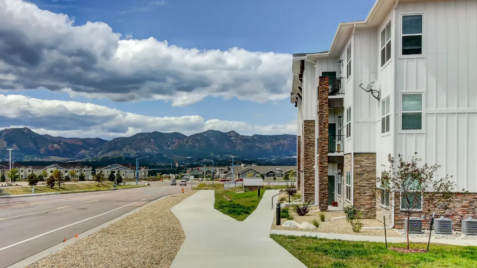 Exterior view of a modern apartment building with a sidewalk, landscaped grounds, and distant mountains.