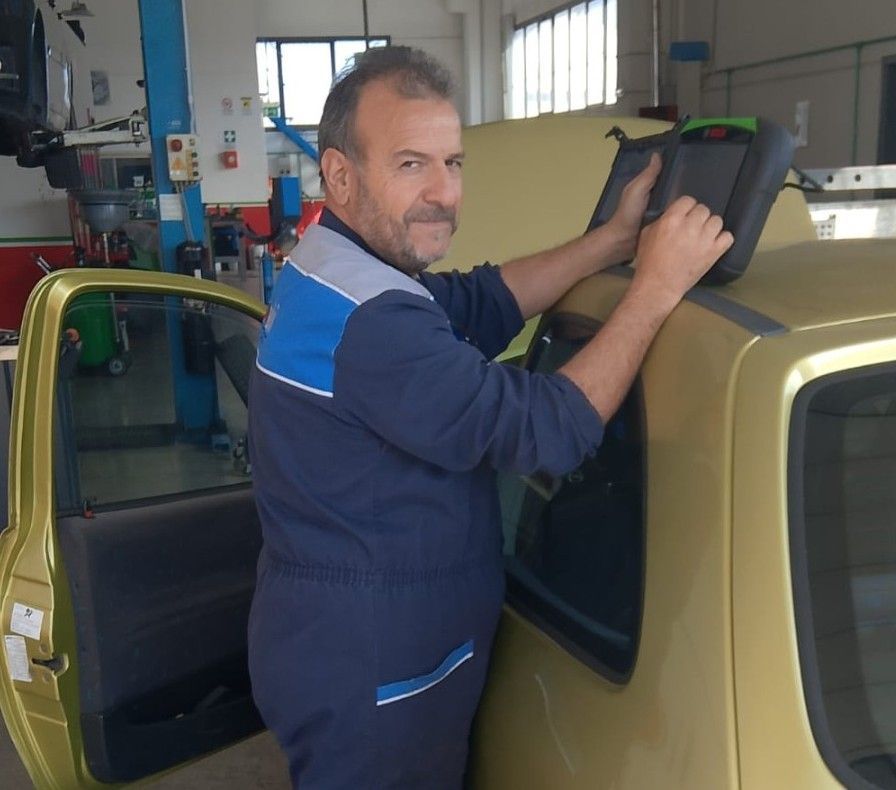 Due meccanici sono in piedi accanto a una macchina di prova in un'officina. Uno indossa un camice verde, l'altro uno blu.