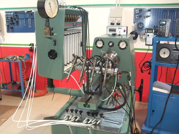 Macchina per il collaudo degli iniettori del motore in un'officina. Verde, con numerosi tubi e manometri, contro una parete rossa/bianca.