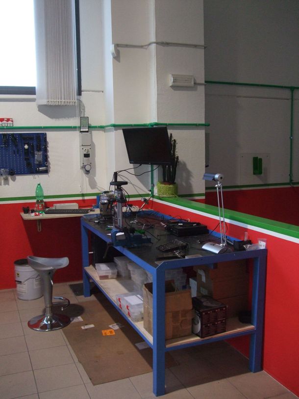 Un laboratorio con un banco da lavoro in metallo blu, attrezzi e uno schermo per computer. Pareti rosse e bianche.