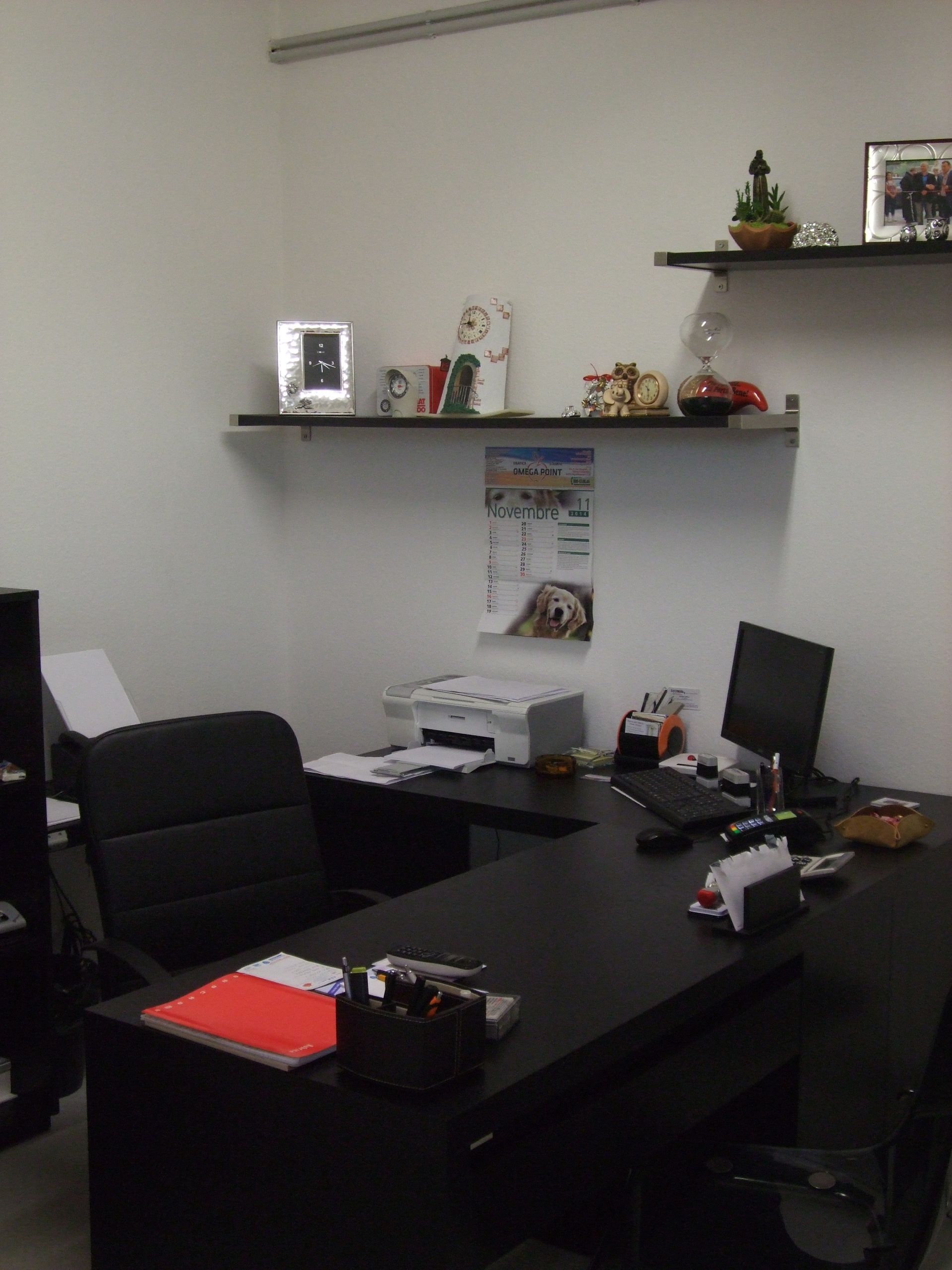 Ufficio con scrivania nera, computer, stampante, scaffali con oggetti e calendario.