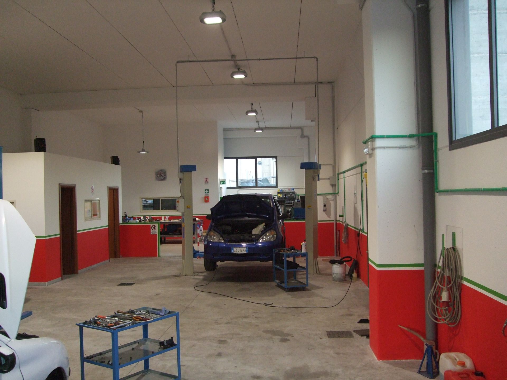 Un'auto all'interno di un garage, sollevata su un paranco per la riparazione. Pareti rosse e bianche, attrezzi e luci.