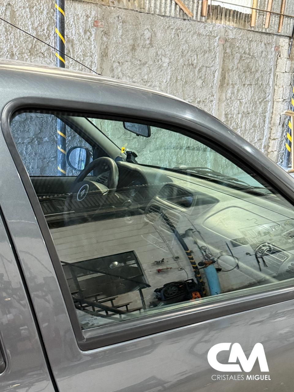 Ventanilla lateral de coche color gris, interior visible: volante, salpicadero, taller reflejado.