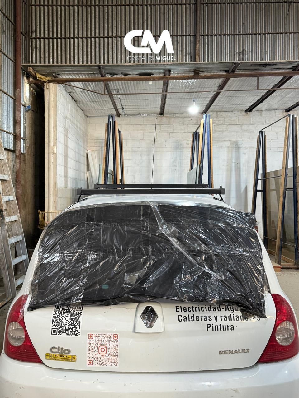 Coche blanco con la ventana trasera cubierta de plástico, bajo un soporte metálico para objetos largos. Interior de tienda.