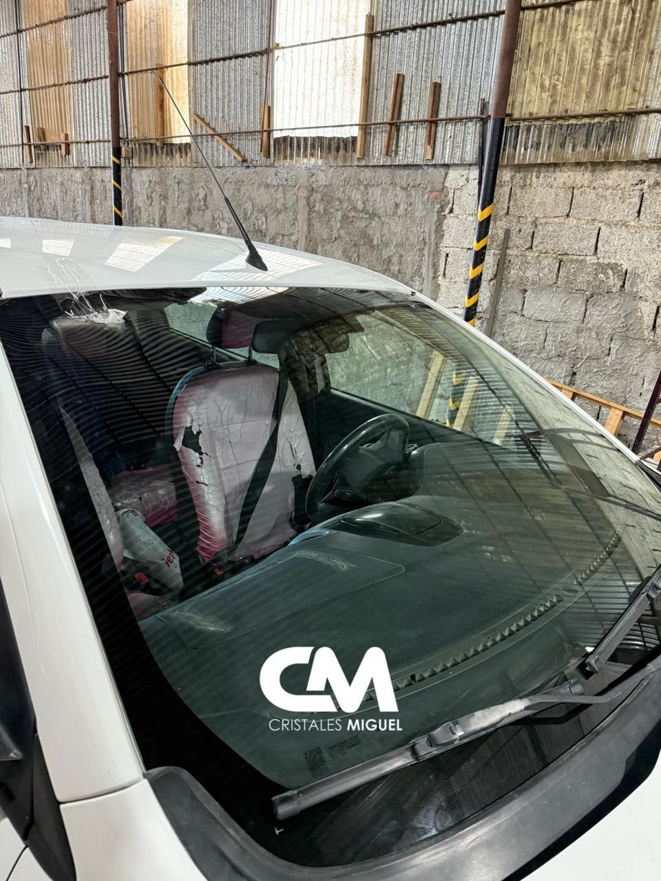 Interior de coche blanco, vista de parabrisas con logotipo, dentro de un edificio con varios materiales visibles.