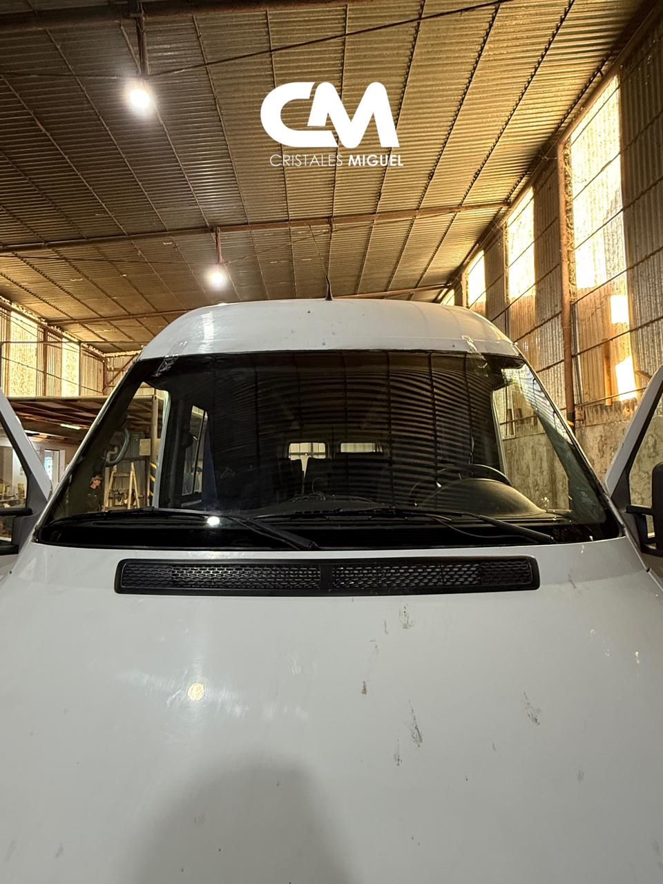 Furgoneta blanca, vista frontal, dentro de un taller con poca luz. Parabrisas protegido por barras metálicas, puertas abiertas.