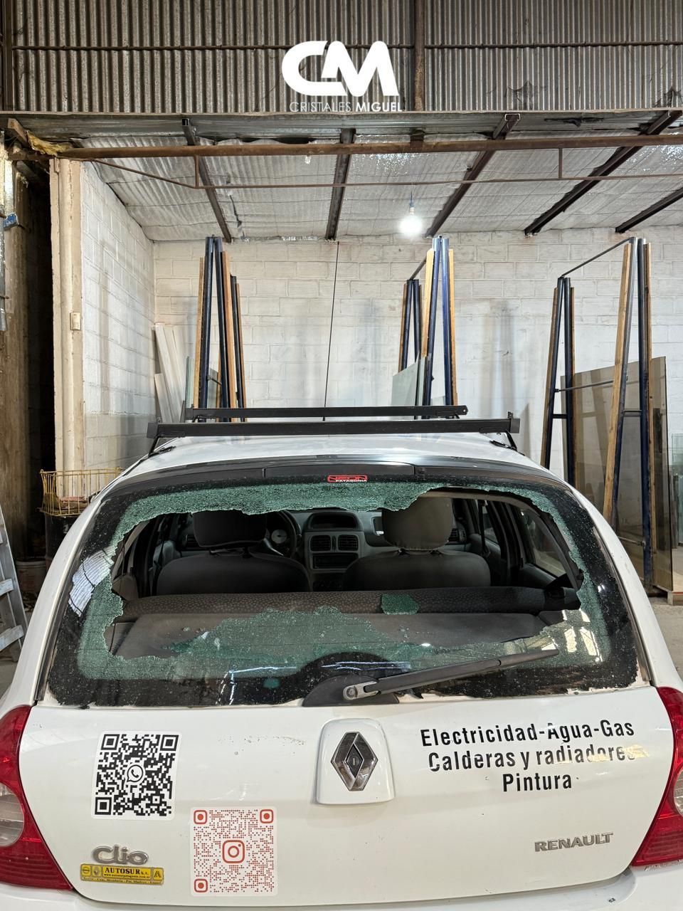 Vista trasera de un coche blanco con la luneta trasera rota, estacionado en un taller. Detrás hay paneles de vidrio apilados.