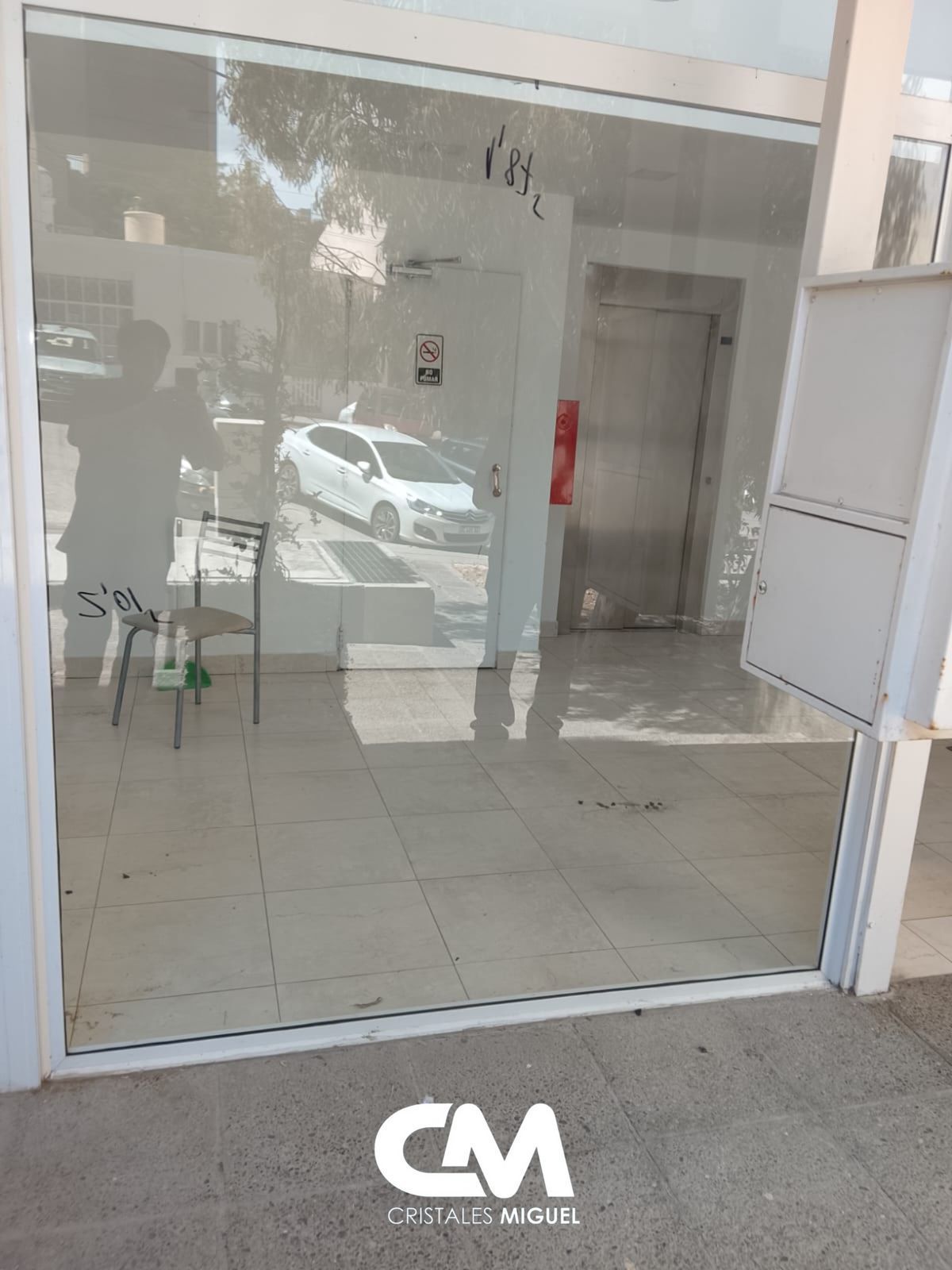 Entrada de cristal con ascensor, coche blanco visible a través de la ventana.
